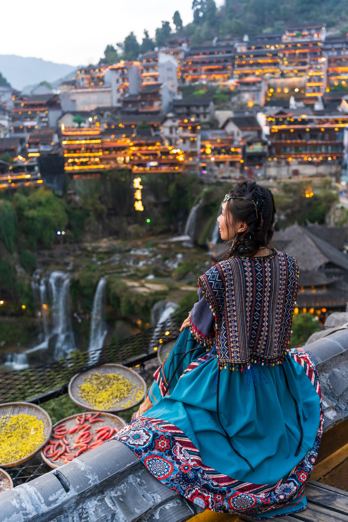 Una mujer china en primer plano sentada de espaldas mirando hacia la ciudad de Lijiang