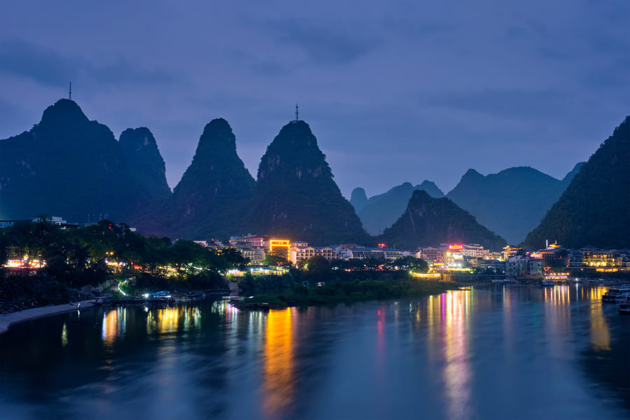 Paisaje nocturno de montañas, una ciudad y en primer plano un lago con el reflejo de las luces en Yangshuo